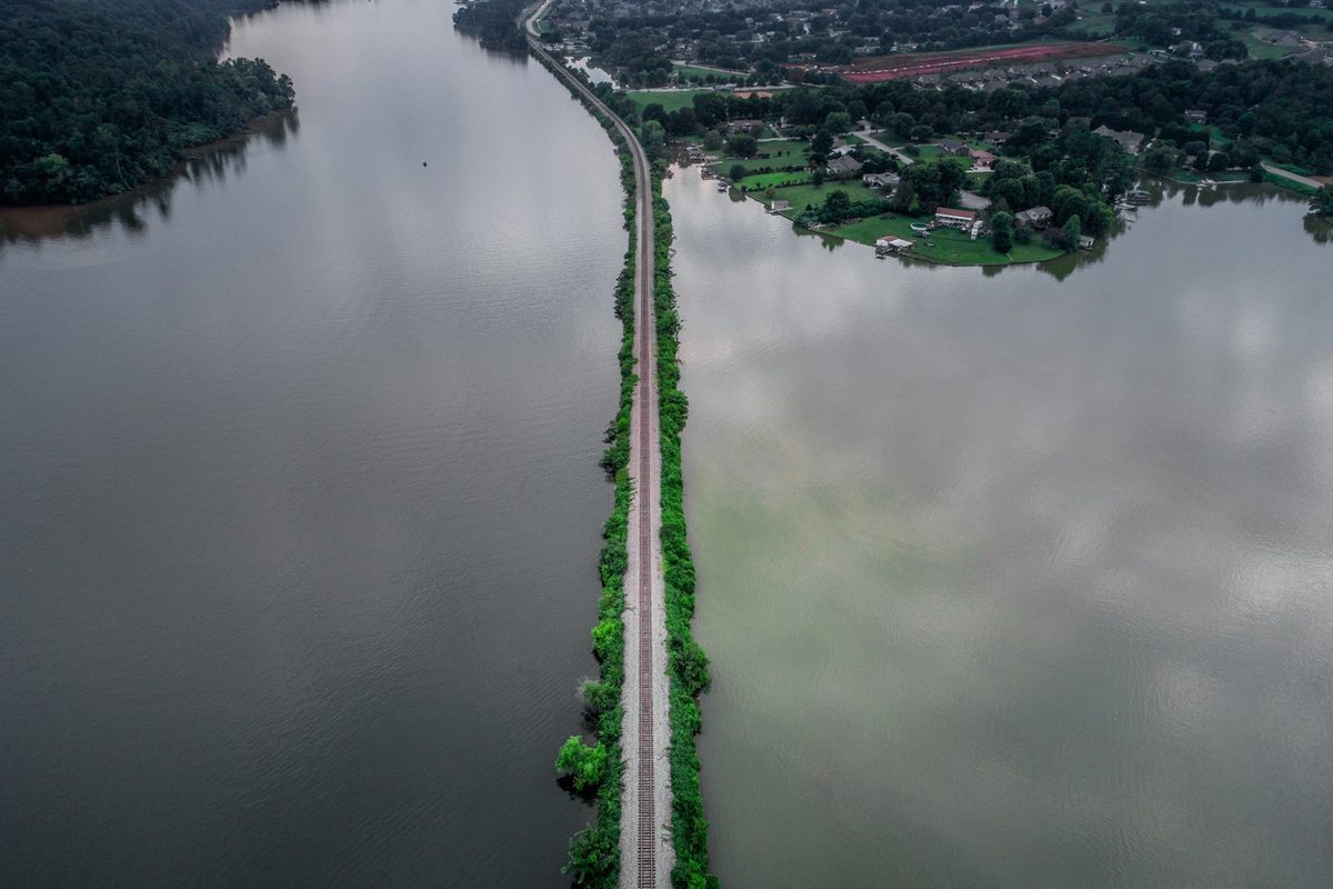 Aerial view of lakefront homes with private docks and a boathouse on a wooded point where a rail causeway crosses the lake.