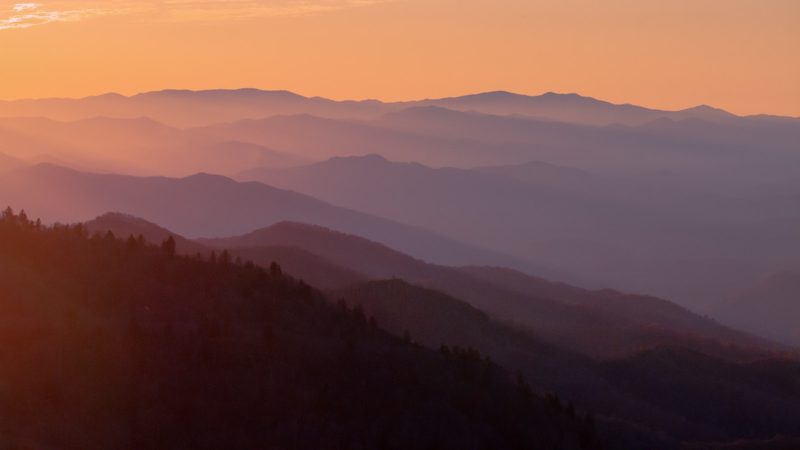 Layered ridges of the Great Smoky Mountains at sunset, deep blue silhouettes fading into haze beneath a wide orange sky.