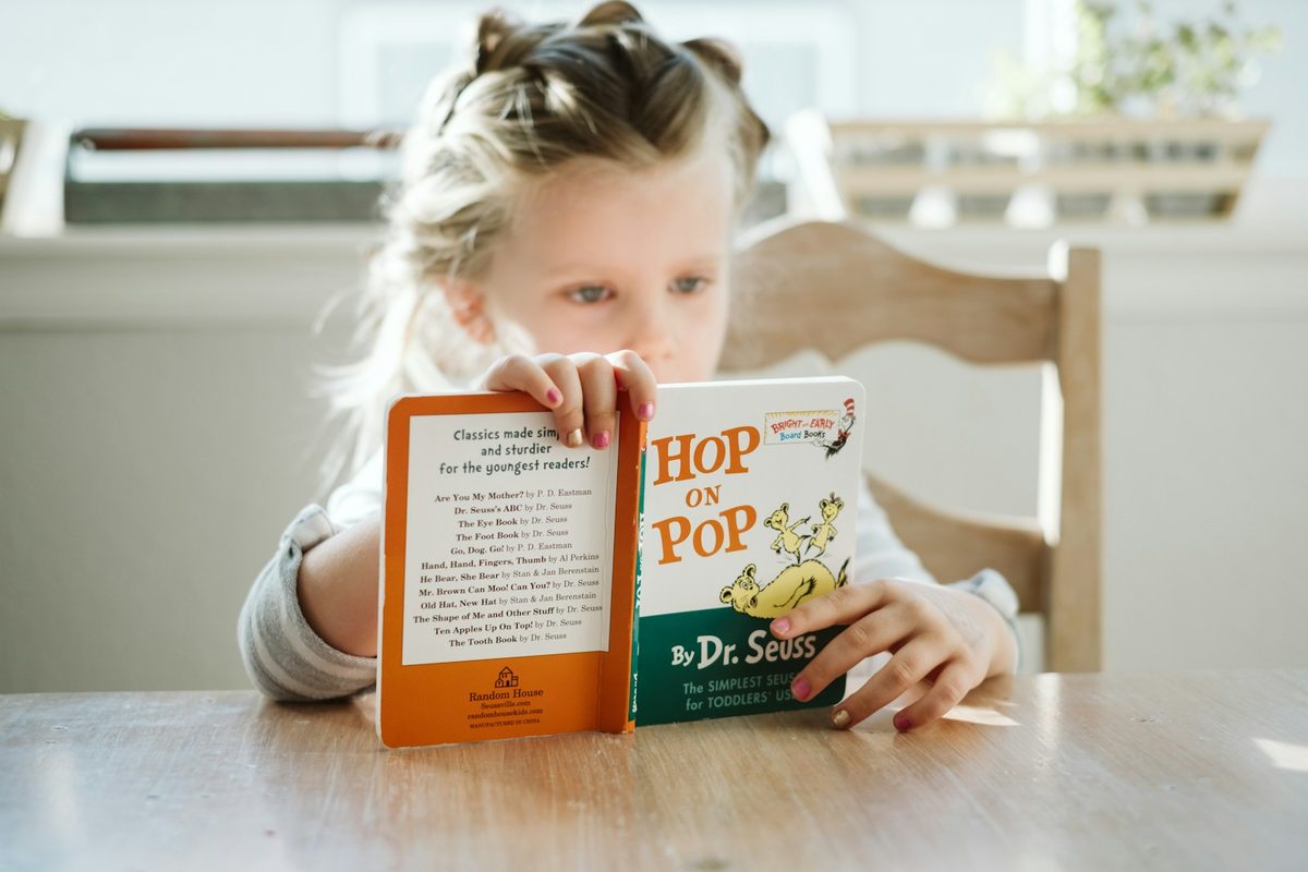 A young girl in a striped shirt sitting at a sunlit wooden kitchen table, holding an open board book up in front of her face.