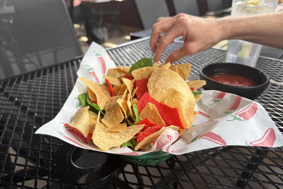 A basket of red, white, and green corn tortilla chips on a Dos Agaves chili-pepper-printed liner with a small ramekin of red salsa, a hand reaching in mid-bite, on a black mesh patio table at Dos Agaves Mexican Grill.