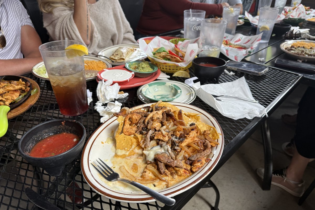 A plate of Dos Agaves nacho fajitas — chicken and steak with onions and a melted cheese sauce over yellow tortillas — on a patio table beside a side of guacamole, tortillas, drinks, and the rest of a group dinner.