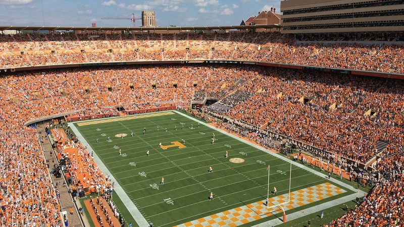 A packed Neyland Stadium on a sunny game day — both decks full of orange-clad fans, the Power T at midfield, and Ayres Hall rising beyond the north rim.