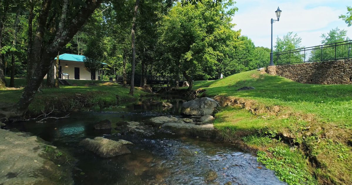 A clear creek with stepping stones running through the manicured grounds of Founders Park, mature shade trees, a small pavilion with a teal metal roof in the background, and a lamppost on a stone retaining wall.