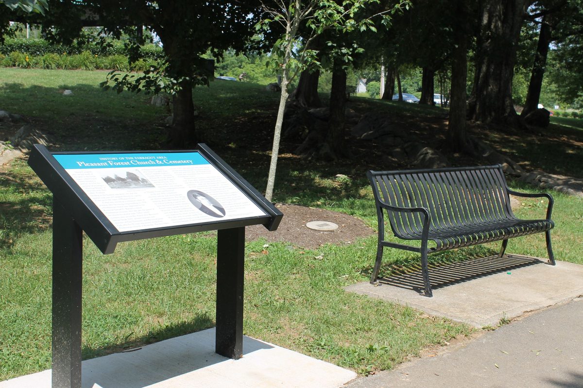 One of the Community Heritage Trail interpretive panels at Founders Park — a black-and-white plaque on a dark metal stand with a black park bench beside it on a small concrete pad, mature trees in the background.