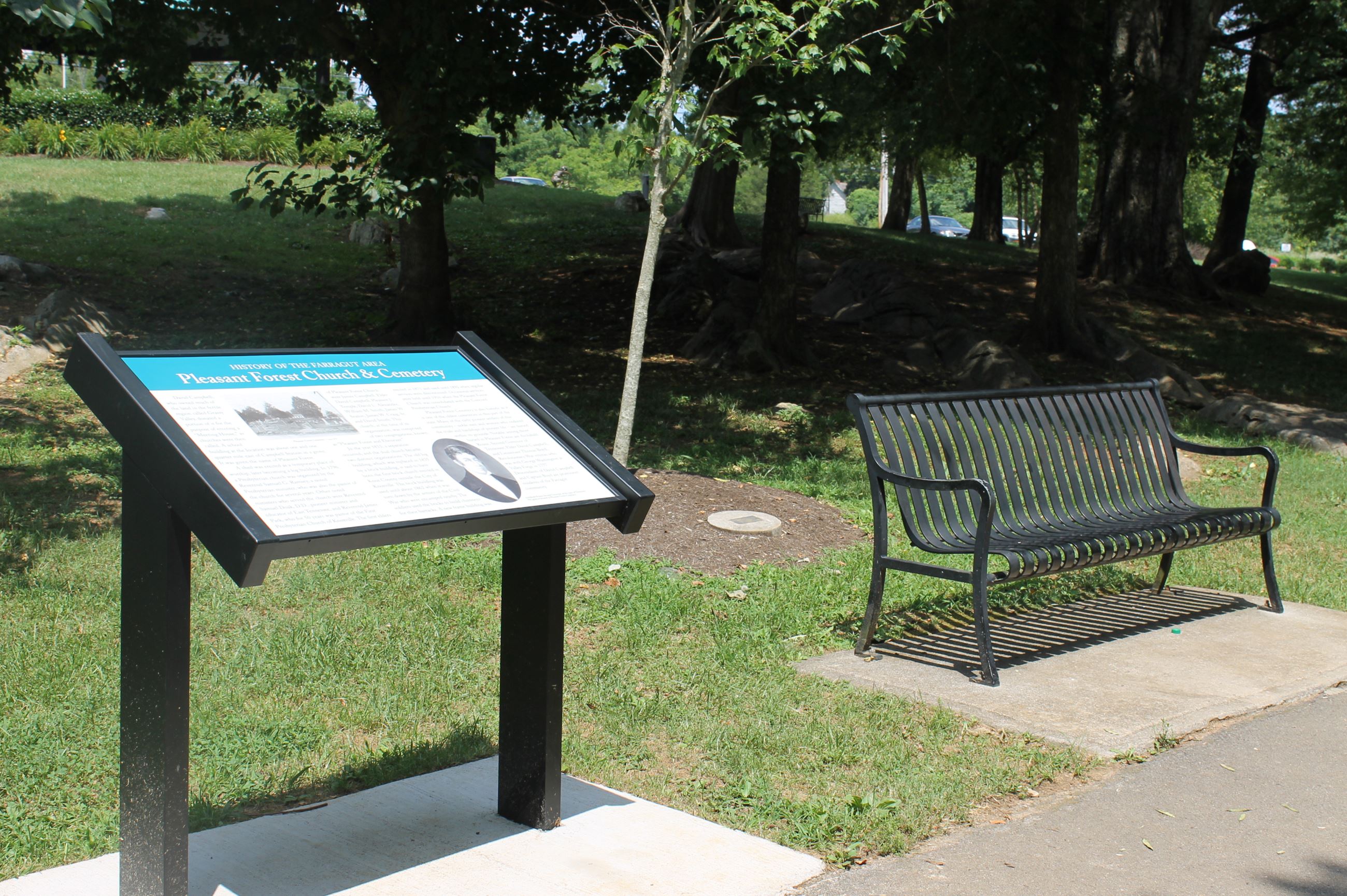One of the Community Heritage Trail interpretive panels at Founders Park — a black-and-white plaque on a dark metal stand with a black park bench beside it on a small concrete pad, mature trees in the background.