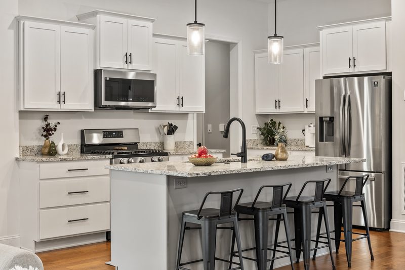 A white kitchen with a granite island, black metal counter stools, glass pendant lights, and a few styled accents on otherwise clear counters.