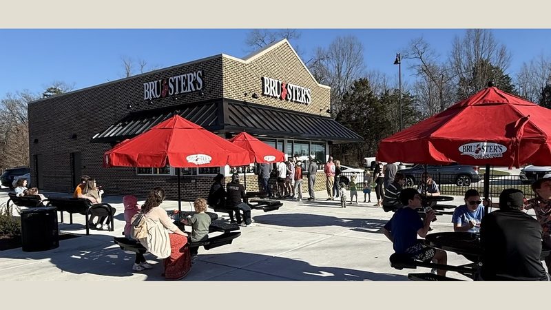 The new Bruster's Real Ice Cream storefront in Turkey Creek (11692 Parkside Drive, Farragut) on a sunny afternoon — families seated at red-umbrella picnic tables, a line of customers at the walk-up window, and the brick-and-board facade with the Bruster's wordmark above the entrance.