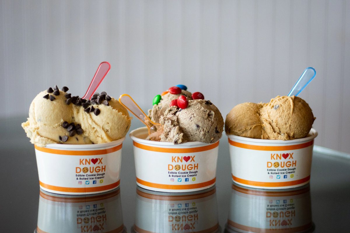Three Knox Dough paper cups lined up on a glass counter — a vanilla scoop with mini chocolate chips, a chocolate scoop topped with M&Ms, and a third caramel-toned scoop — each cup branded in the shop's orange-and-white wordmark, photographed in soft natural light at the Kingston Pike location.