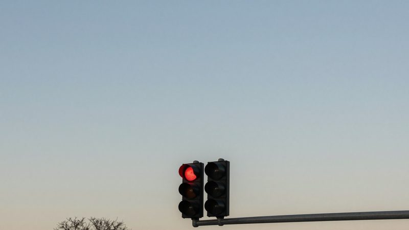 A new traffic signal glowing red on a fresh galvanized mast arm against a soft gold-and-blue dusk sky, with a bare hardwood tree silhouetted at the lower edge of a West Farragut street.