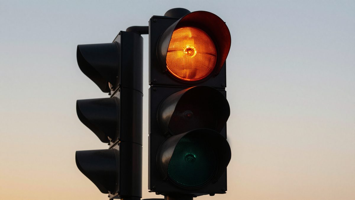 An extreme close-up of a traffic signal head with the amber lamp glowing warmly against a soft gold-and-blue dusk sky, the kind of flashing-yellow phase a new signal runs in before it begins its full cycle.