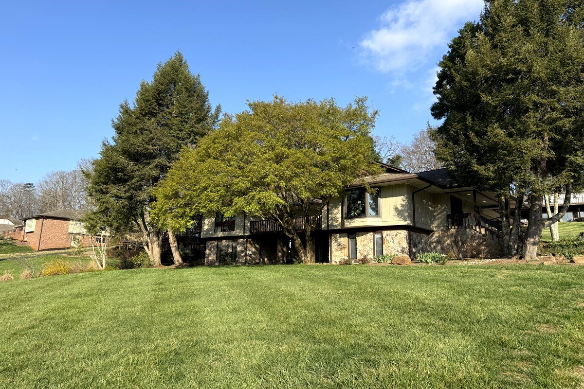 A split-level home in Fox Den, framed by mature dogwoods and evergreens, set behind a wide green spring lawn under a clear blue Tennessee sky.