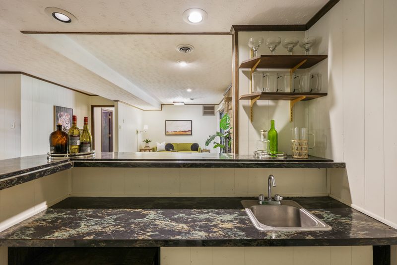 Stone wet bar with dark counter and sink, looking out over the lower-level rec room